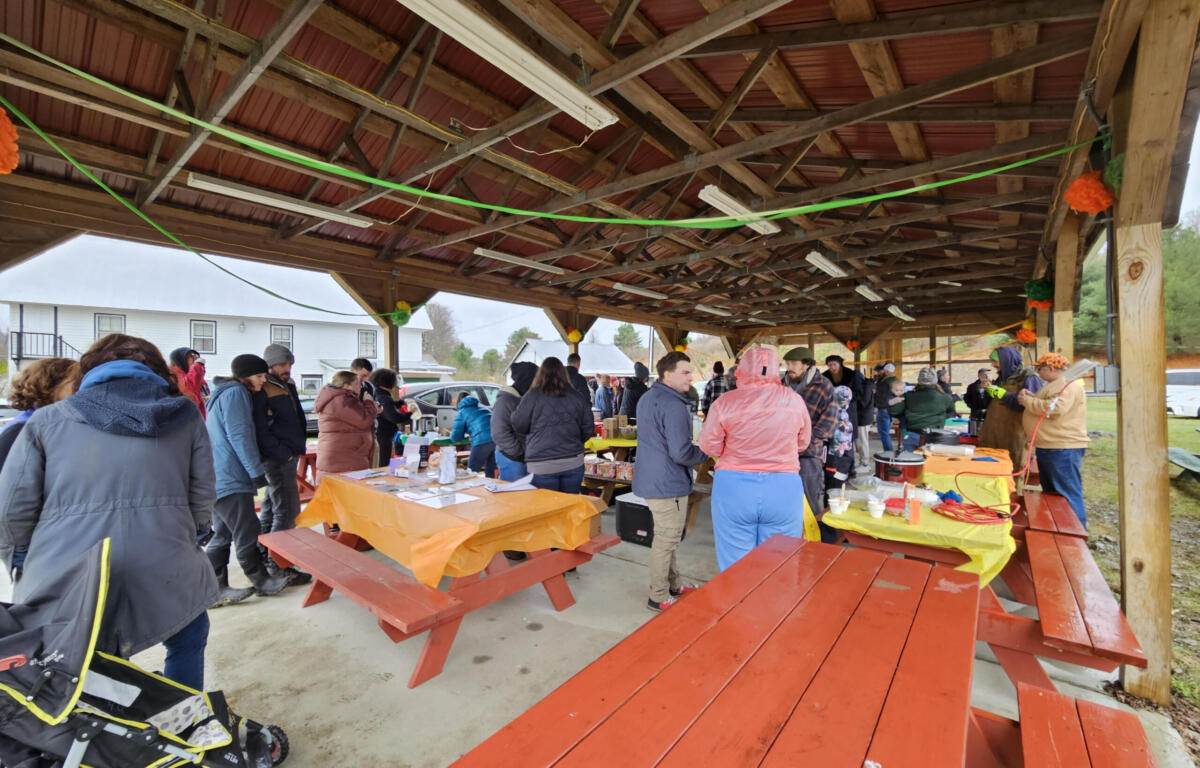People gather under a wooden pavilion at an outdoor community event, around orange-clothed tables sharing food and conversations.