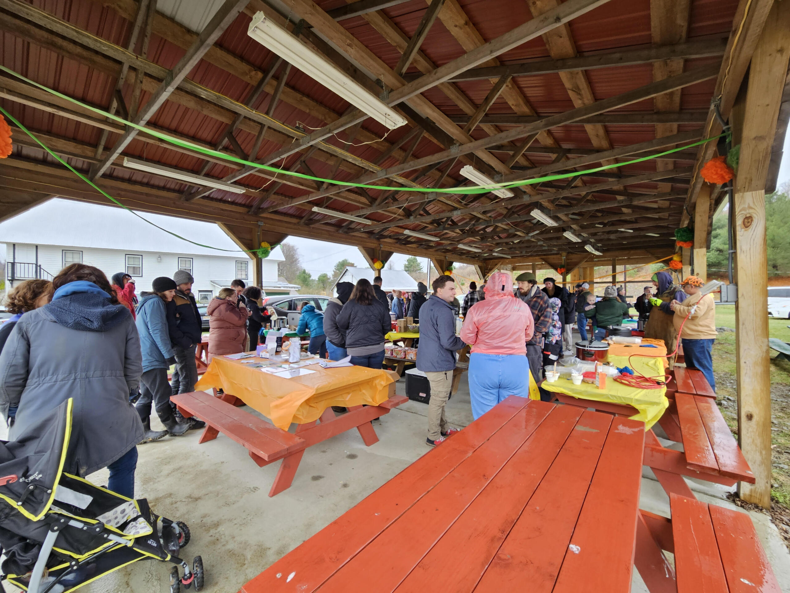 People gather under a wooden pavilion at an outdoor community event, around orange-clothed tables sharing food and conversations.