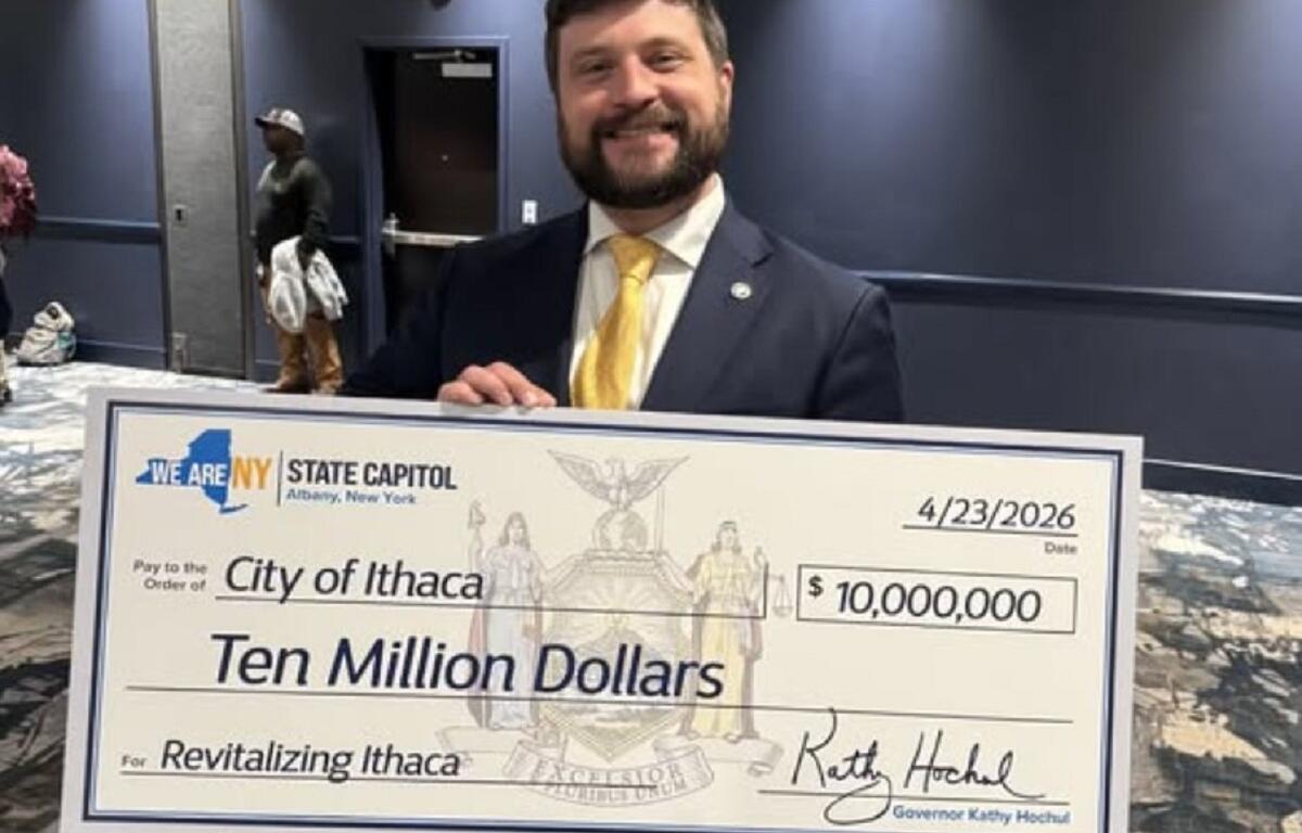 Smiling man in a dark suit and yellow tie holds a large ceremonial check reading Ten Million Dollars for Revitalizing Ithaca at the State Capitol, dated 4/23/2026 and showing 'We Are NY' logo.