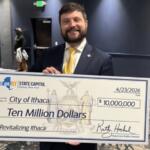 Smiling man in a dark suit and yellow tie holds a large ceremonial check reading Ten Million Dollars for Revitalizing Ithaca at the State Capitol, dated 4/23/2026 and showing 'We Are NY' logo.