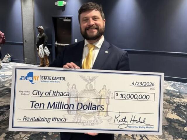 Smiling man in a dark suit and yellow tie holds a large ceremonial check reading Ten Million Dollars for Revitalizing Ithaca at the State Capitol, dated 4/23/2026 and showing 'We Are NY' logo.