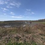 Group of people in safety vests near trucks in a muddy field on a clear day.
