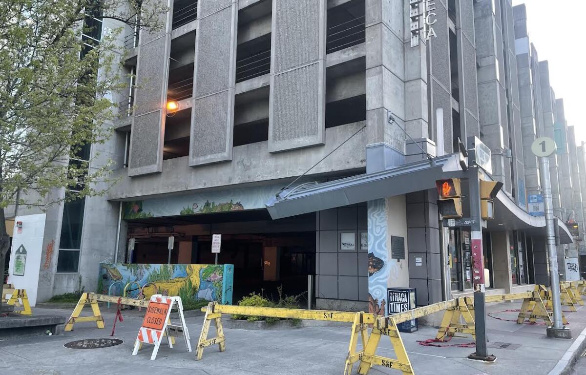 Street view of a concrete multi-story building with a blocked ground-floor entrance, yellow barricades, and an orange sidewalk-closure sign in front.