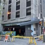 Street view of a concrete multi-story building with a blocked ground-floor entrance, yellow barricades, and an orange sidewalk-closure sign in front.