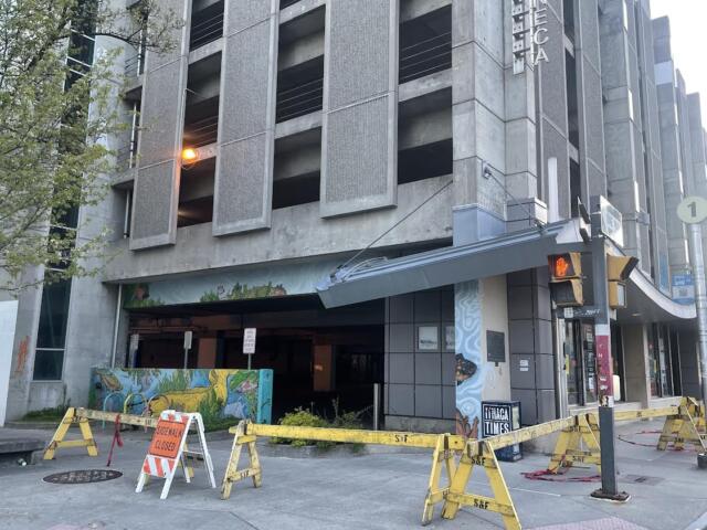 Street view of a concrete multi-story building with a blocked ground-floor entrance, yellow barricades, and an orange sidewalk-closure sign in front.