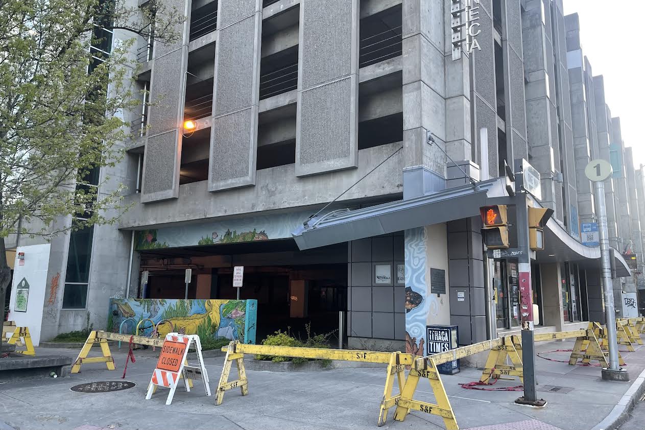 Street view of a concrete multi-story building with a blocked ground-floor entrance, yellow barricades, and an orange sidewalk-closure sign in front.