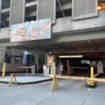 Entrance to a parking garage with yellow barriers and a horizontal height bar, concrete building facade above, and a mural on the wall.