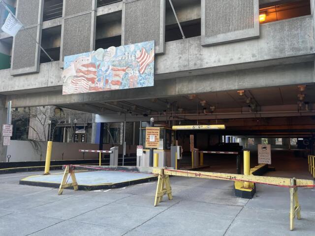 Entrance to a parking garage with yellow barriers and a horizontal height bar, concrete building facade above, and a mural on the wall.