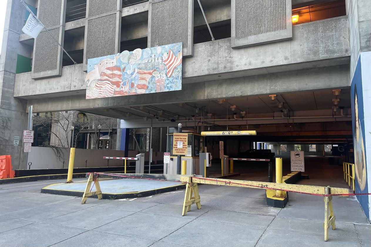 Entrance to a parking garage with yellow barriers and a horizontal height bar, concrete building facade above, and a mural on the wall.