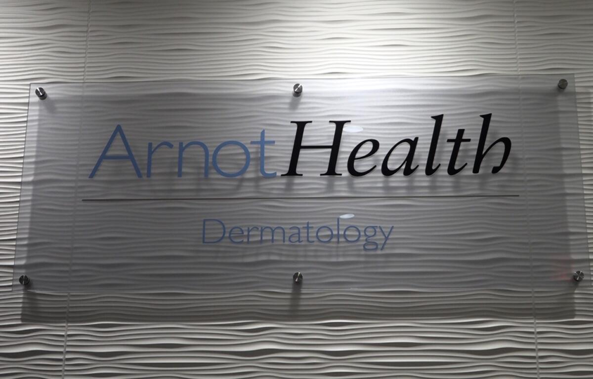 Sign reading 'ArnotHealth Dermatology' mounted on a frosted glass panel with a wavy textured wall background.