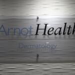 Sign reading 'ArnotHealth Dermatology' mounted on a frosted glass panel with a wavy textured wall background.