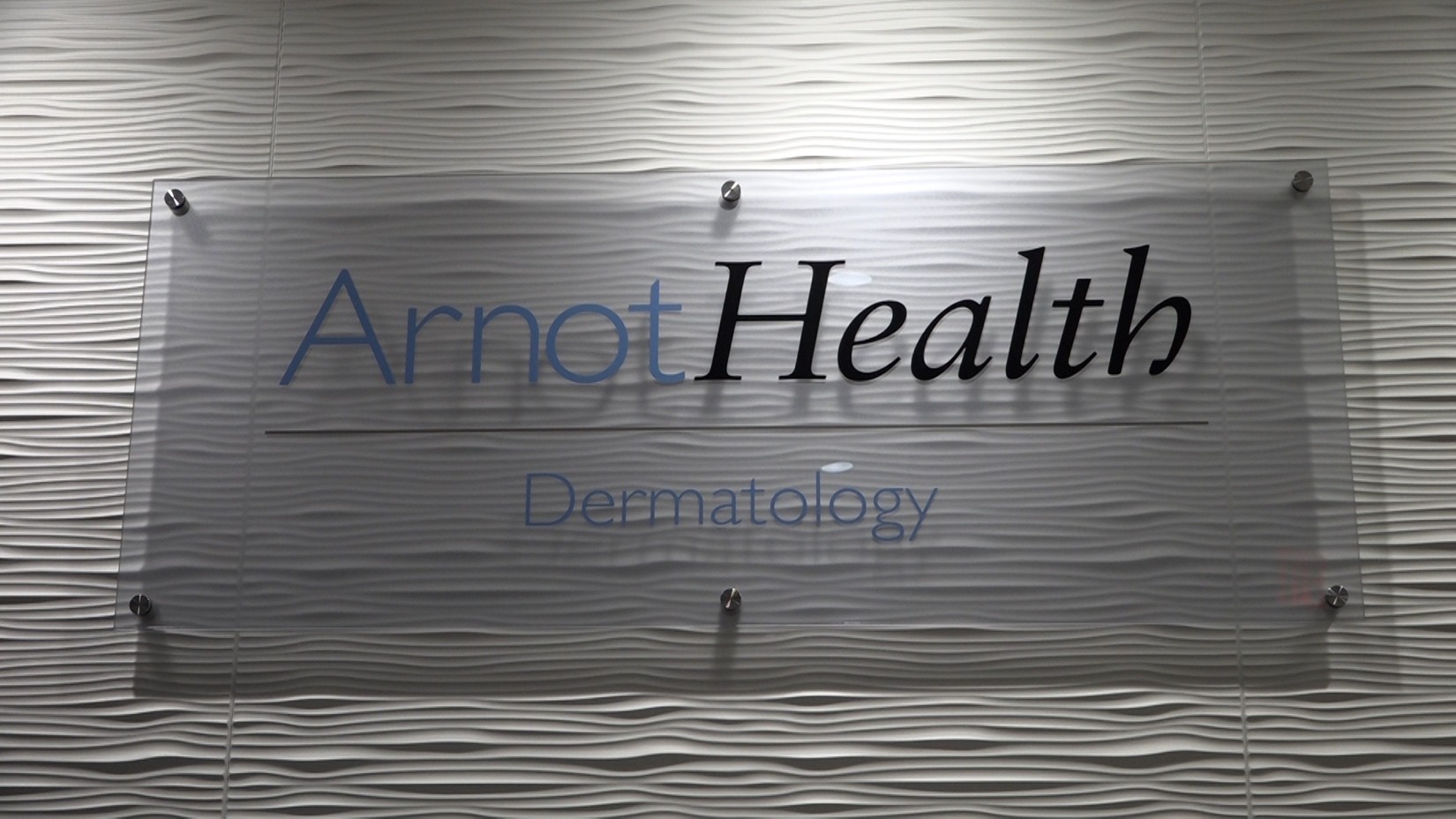 Sign reading 'ArnotHealth Dermatology' mounted on a frosted glass panel with a wavy textured wall background.