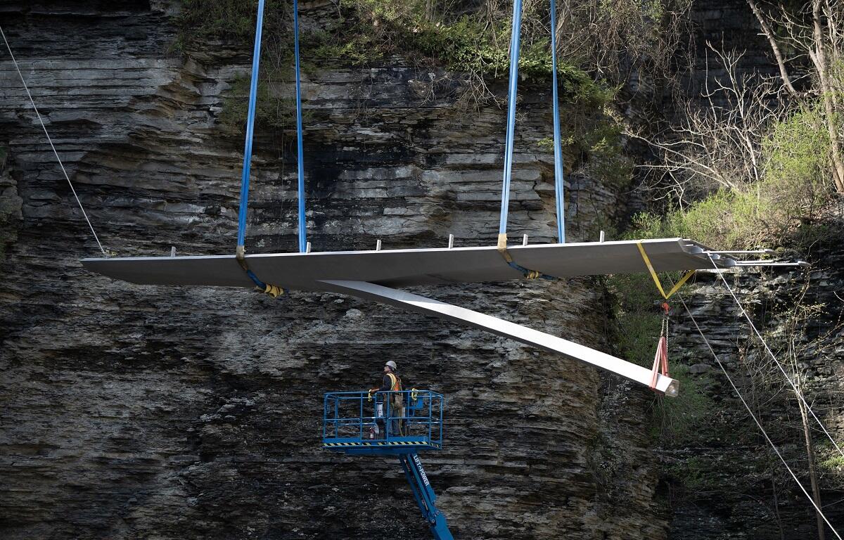 Large flat wing‑like panel being hoisted by blue straps over a rocky cliff, with a worker standing in a blue lift basket nearby.