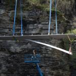 Large flat wing‑like panel being hoisted by blue straps over a rocky cliff, with a worker standing in a blue lift basket nearby.