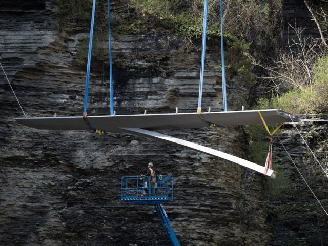 Large flat wing‑like panel being hoisted by blue straps over a rocky cliff, with a worker standing in a blue lift basket nearby.