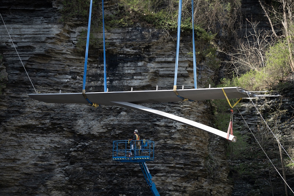 Large flat wing‑like panel being hoisted by blue straps over a rocky cliff, with a worker standing in a blue lift basket nearby.