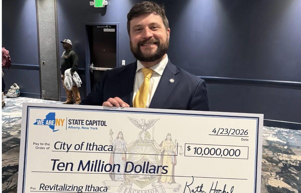 Man in a dark suit and yellow tie smiling as he holds a large ceremonial check reading 'Ten Million Dollars' to 'Revitalizing Ithaca' with date 4/23/2026 and a 'City of Ithaca' designation. The check is branded with 'We Are NY State Capitol' and features a crest in the center.