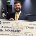 Man in a dark suit and yellow tie smiling as he holds a large ceremonial check reading 'Ten Million Dollars' to 'Revitalizing Ithaca' with date 4/23/2026 and a 'City of Ithaca' designation. The check is branded with 'We Are NY State Capitol' and features a crest in the center.