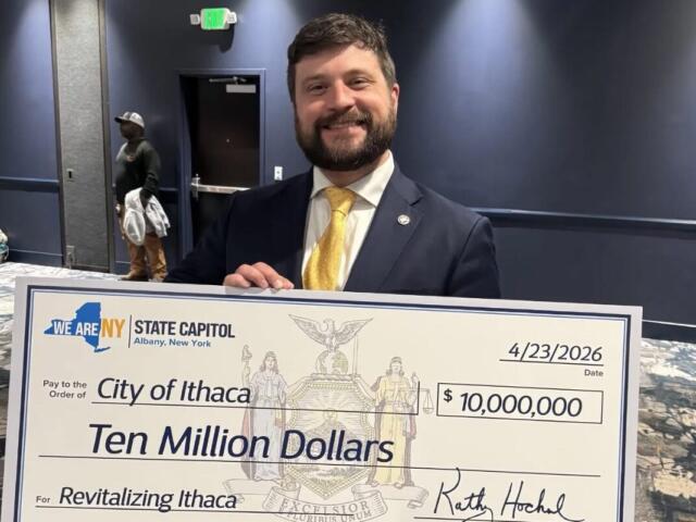 Man in a dark suit and yellow tie smiling as he holds a large ceremonial check reading 'Ten Million Dollars' to 'Revitalizing Ithaca' with date 4/23/2026 and a 'City of Ithaca' designation. The check is branded with 'We Are NY State Capitol' and features a crest in the center.