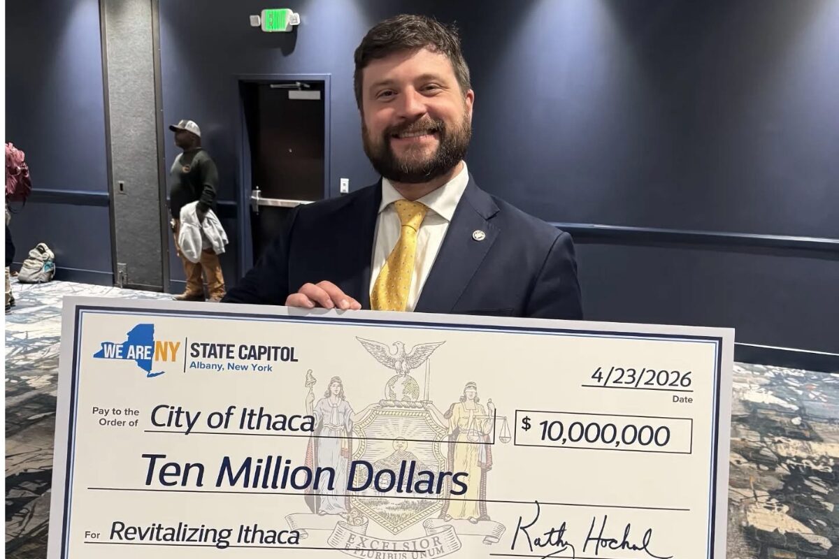 Man in a dark suit and yellow tie smiling as he holds a large ceremonial check reading 'Ten Million Dollars' to 'Revitalizing Ithaca' with date 4/23/2026 and a 'City of Ithaca' designation. The check is branded with 'We Are NY State Capitol' and features a crest in the center.