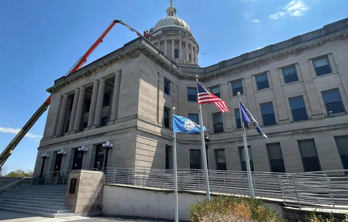 Neoclassical government building with a dome, flagpoles, and a construction lift extending over the roof against a blue sky.