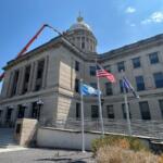 Neoclassical government building with a dome, flagpoles, and a construction lift extending over the roof against a blue sky.