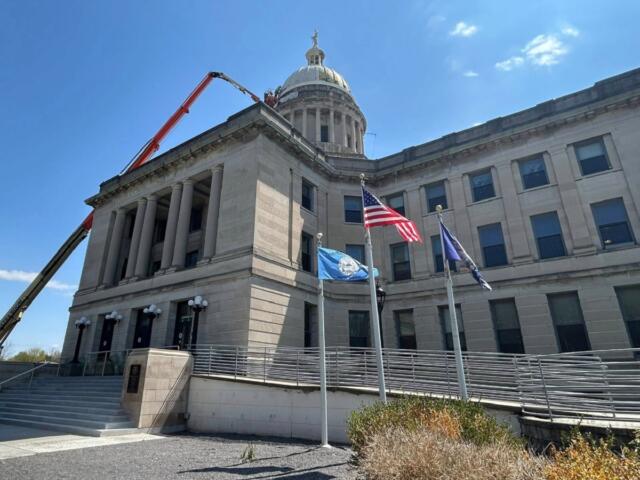 Neoclassical government building with a dome, flagpoles, and a construction lift extending over the roof against a blue sky.