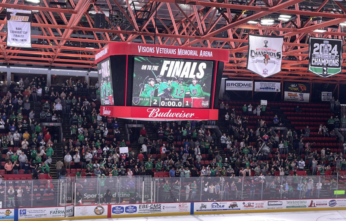 Crowd in green jerseys watches a hockey game as the scoreboard reads FINALS and a 2-1 score. Admin banners hang from the red ceiling.