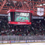 Crowd in green jerseys watches a hockey game as the scoreboard reads FINALS and a 2-1 score. Admin banners hang from the red ceiling.