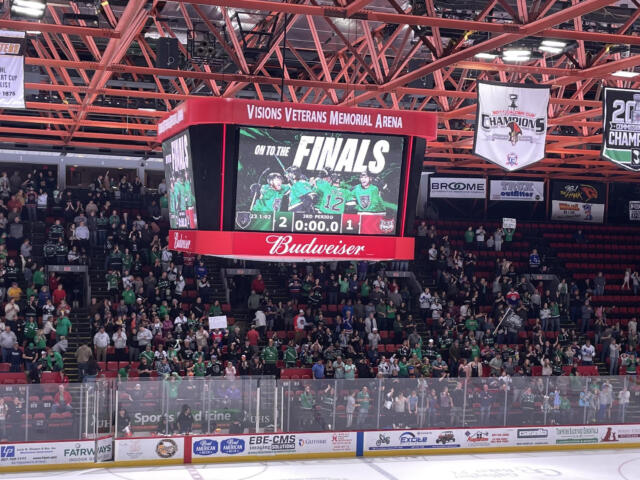 Crowd in green jerseys watches a hockey game as the scoreboard reads FINALS and a 2-1 score. Admin banners hang from the red ceiling.