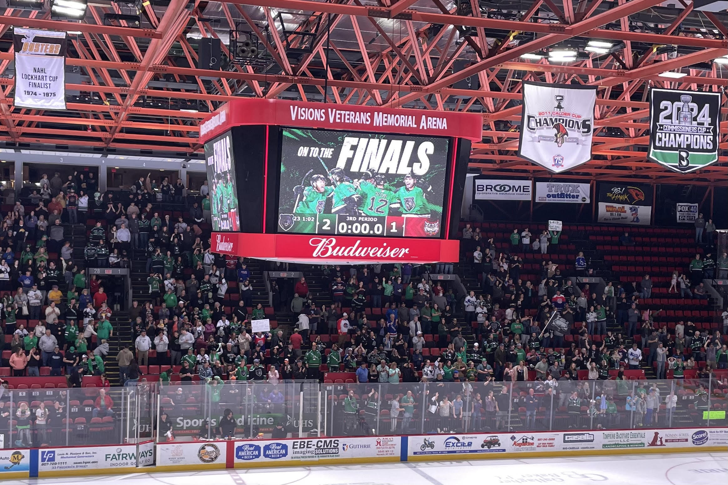 Crowd in green jerseys watches a hockey game as the scoreboard reads FINALS and a 2-1 score. Admin banners hang from the red ceiling.
