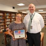Elizabeth Kozlowski holds a framed certificate of achievement with a smiling man in a library.