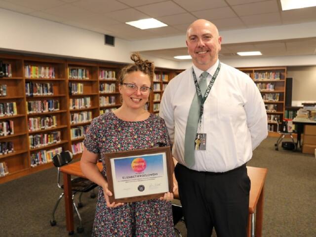 Elizabeth Kozlowski holds a framed certificate of achievement with a smiling man in a library.