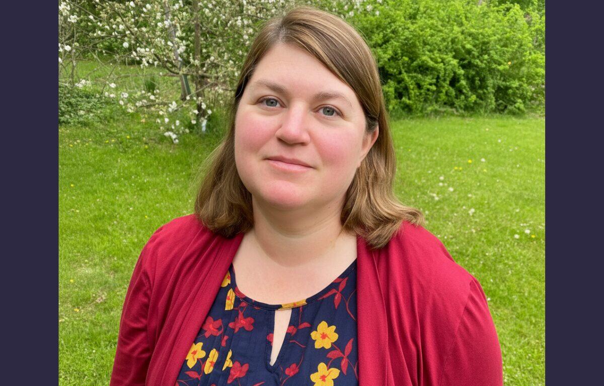 Portrait of a woman with light brown hair standing in a grassy park, wearing a red cardigan over a navy floral blouse.