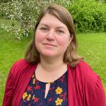 Portrait of a woman with light brown hair standing in a grassy park, wearing a red cardigan over a navy floral blouse.