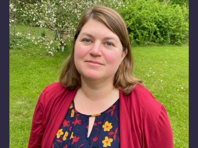 Portrait of a woman with light brown hair standing in a grassy park, wearing a red cardigan over a navy floral blouse.