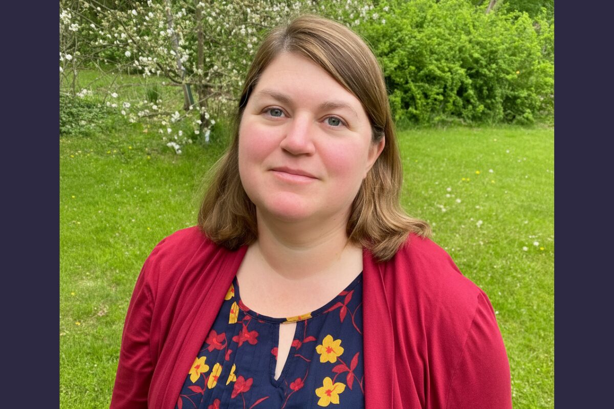 Portrait of a woman with light brown hair standing in a grassy park, wearing a red cardigan over a navy floral blouse.