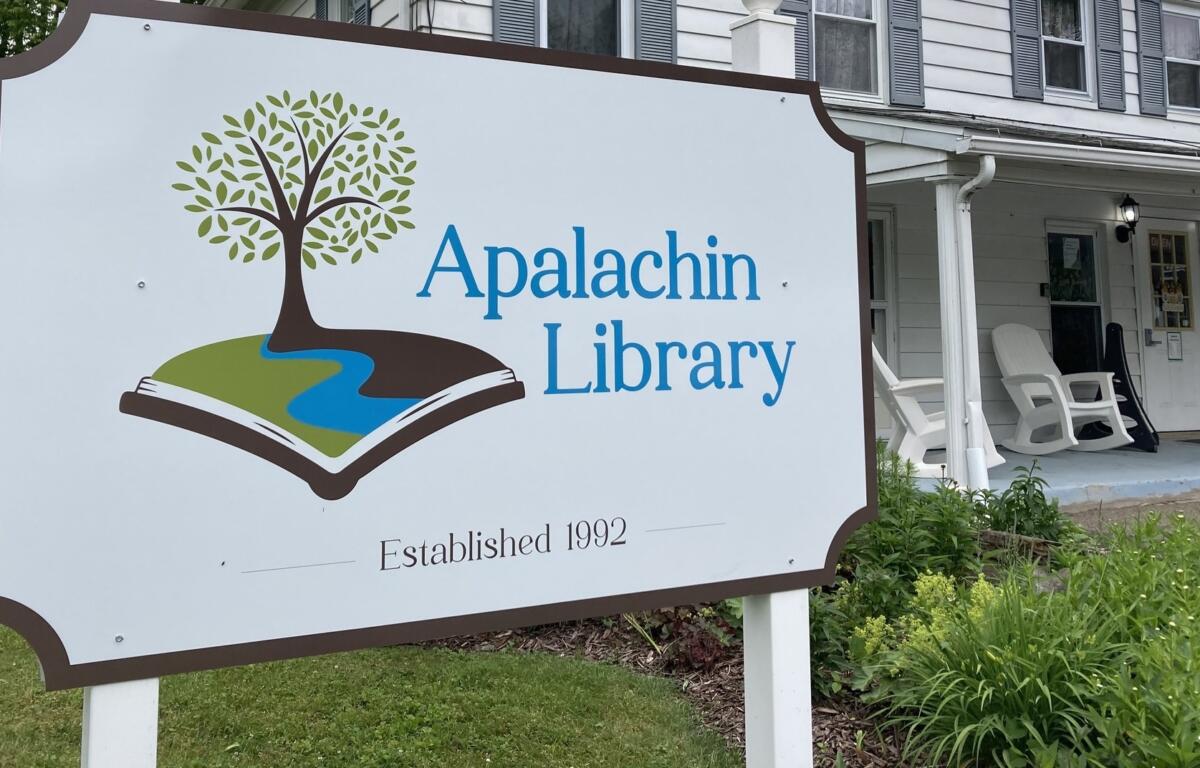 Sign for Apalachin Library featuring a tree growing from an open book and 'Established 1992' on a lawn scene.