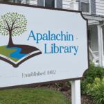 Sign for Apalachin Library featuring a tree growing from an open book and 'Established 1992' on a lawn scene.