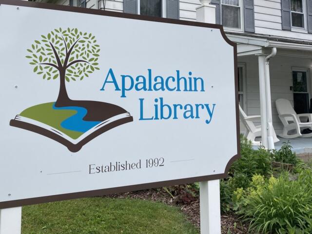 Sign for Apalachin Library featuring a tree growing from an open book and 'Established 1992' on a lawn scene.