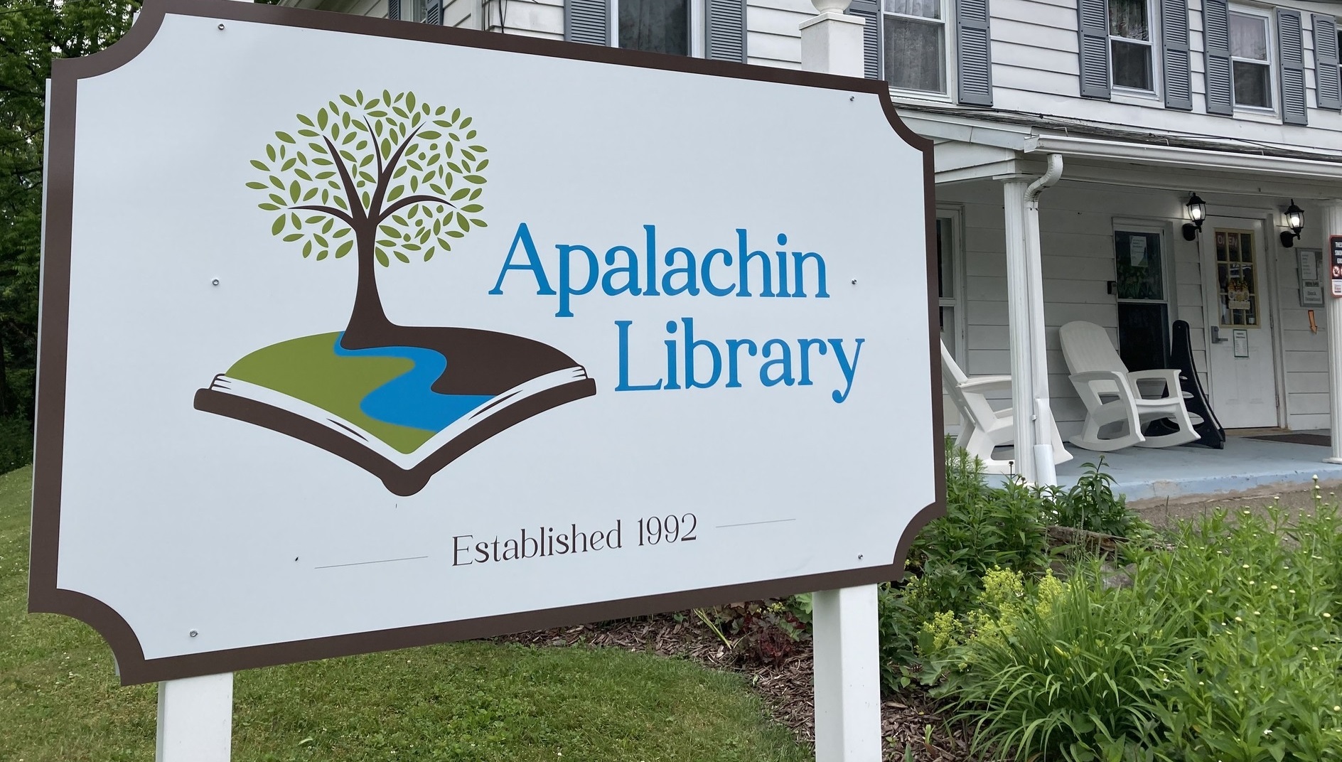 Sign for Apalachin Library featuring a tree growing from an open book and 'Established 1992' on a lawn scene.