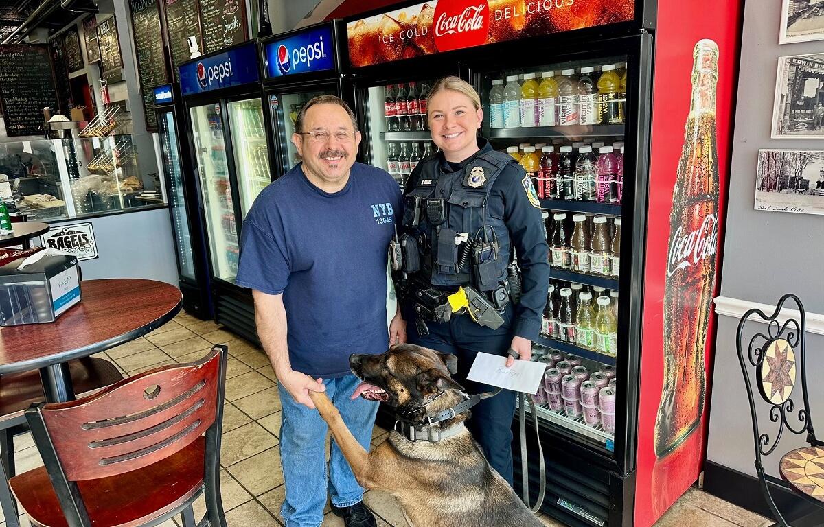 Police officer in uniform and a man pose with a German shepherd K-9 in a café, standing by a Coca-Cola cooler and soda fridge, the dog offering its paw.