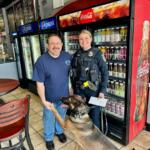 Police officer in uniform and a man pose with a German shepherd K-9 in a café, standing by a Coca-Cola cooler and soda fridge, the dog offering its paw.