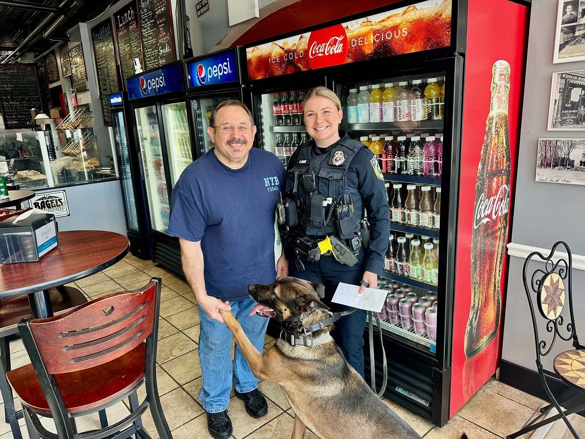 Police officer in uniform and a man pose with a German shepherd K-9 in a café, standing by a Coca-Cola cooler and soda fridge, the dog offering its paw.