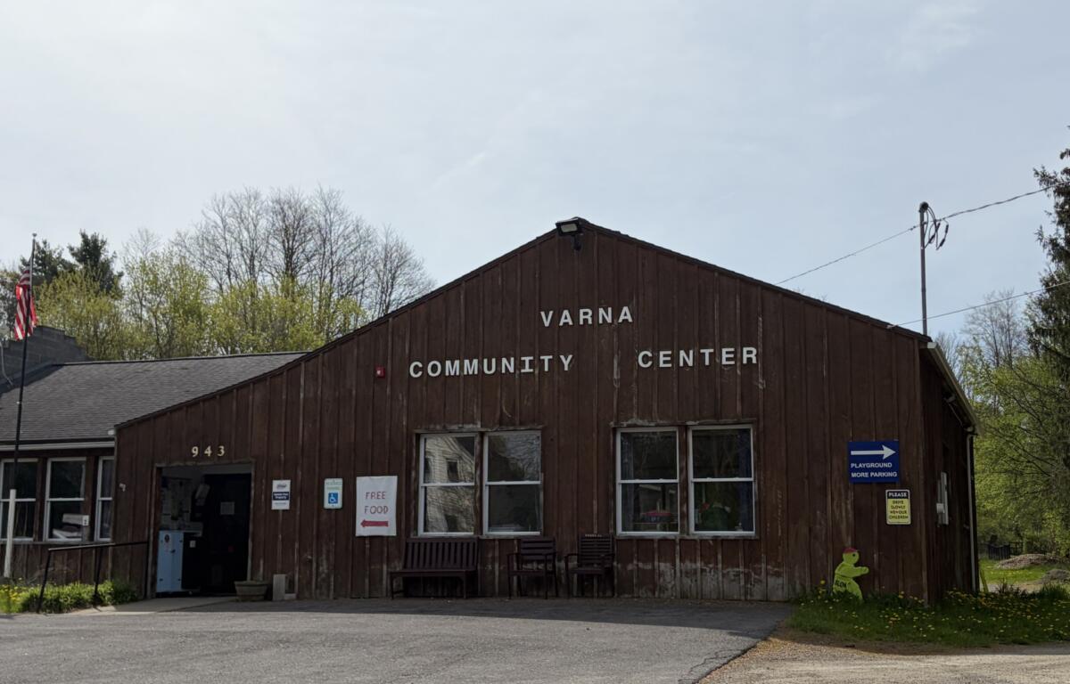 Front of Varna Community Center: brown wooden building with 'VARNA COMMUNITY CENTER' signage, entrance doors, and parking area in foreground, flags and signs nearby.