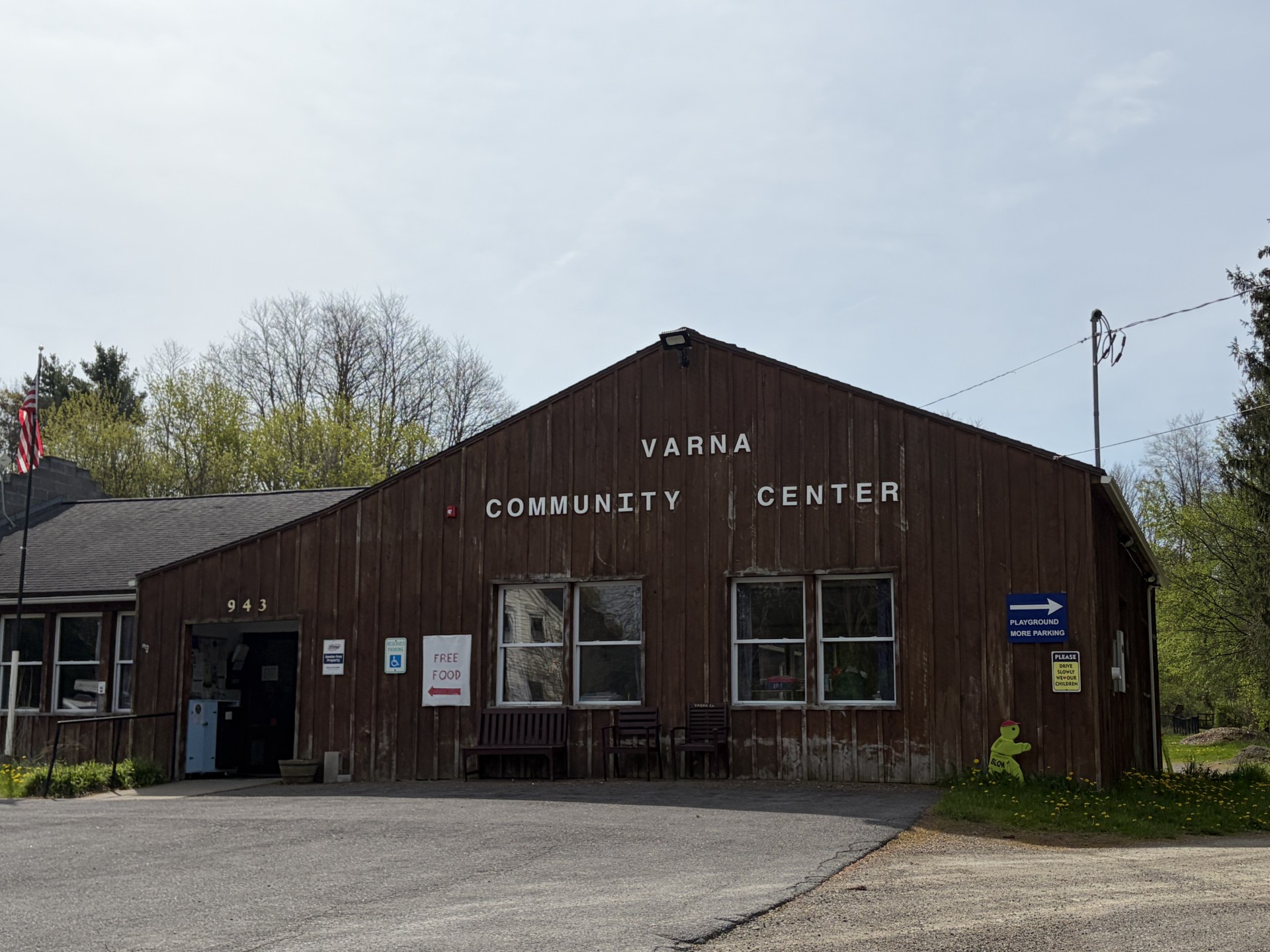 Front of Varna Community Center: brown wooden building with 'VARNA COMMUNITY CENTER' signage, entrance doors, and parking area in foreground, flags and signs nearby.