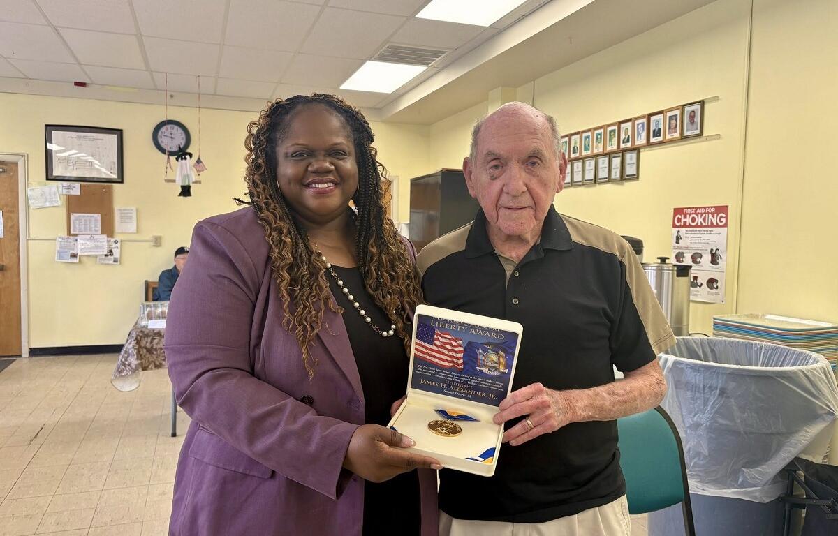 Smiling woman in a purple blazer presents a Liberty Award medal to an elderly man inside a yellow-walled community room.