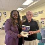 Smiling woman in a purple blazer presents a Liberty Award medal to an elderly man inside a yellow-walled community room.