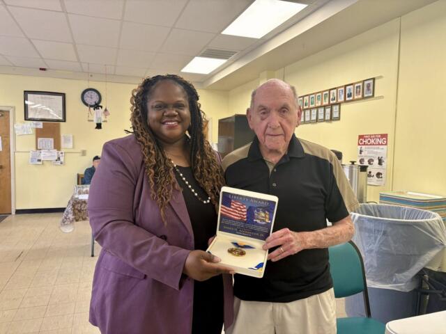 Smiling woman in a purple blazer presents a Liberty Award medal to an elderly man inside a yellow-walled community room.
