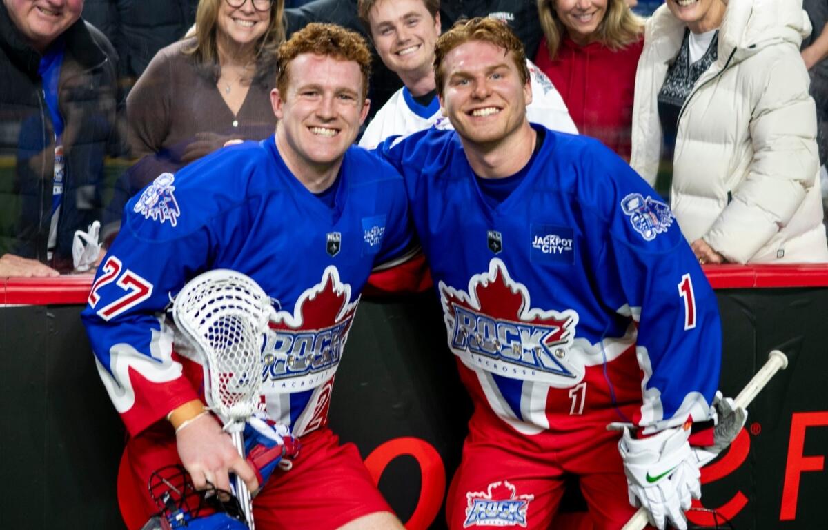 Two hockey players in blue jerseys pose and smile for the camera near the rink boards, fans behind them celebrate.